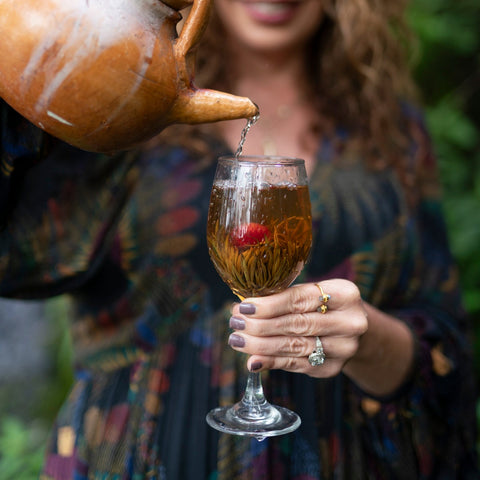 A person pours Magic Hour's Divine Tea: Handmade Flowering Tea from Sri Lanka from a rustic clay teapot into a transparent glass filled with flowering tea and a raspberry. The person wears a dark floral dress and has manicured nails painted in a lavender shade, along with rings on their fingers.