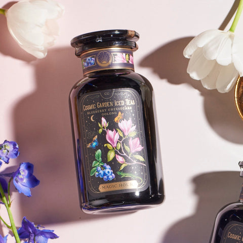 A dark-colored, elegantly designed tea canister labeled "Magic Hour - Blueberry Cheesecake Oolong Iced Tea" is surrounded by white tulips and blue flowers. The canister, featuring a whimsical illustration of flowers and organic blueberries, promises a delightful iced tea experience.