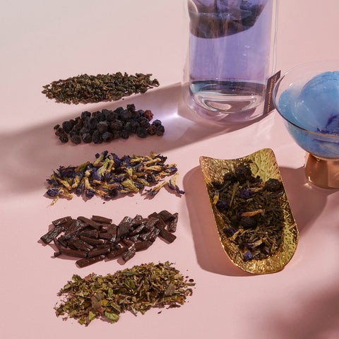 An assortment of dried herbs and organic blueberries arranged in small piles on a light pink surface. Next to them is a glass filled with Magic Hour Blueberry Cheesecake Oolong Iced Tea and a golden dish containing a herb blend. A blue object in a glass container is also visible.