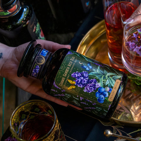 A hand holds a jar labeled "Magic Hour's Blueberry Lavender Mint: Cosmic Garden Iced Tea" with an intricate design featuring blueberries and purple flowers. The jar, brimming with fragrant loose leaf tea, is held over a gold tray surrounded by ornate glasses of beverages. The scene is dimly lit, creating a cozy and enchanting atmosphere.