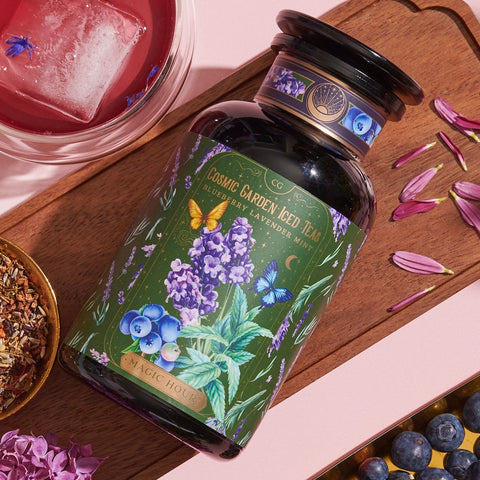 A jar of Blueberry Lavender Mint: Cosmic Garden Iced Tea sits on a wooden tray. The jar features floral and berry designs. Surrounding the jar are petals, loose leaf tea mix, blueberries, and a glass of iced tea with a pinkish hue. This Magic Hour product promises an enchanting sip.