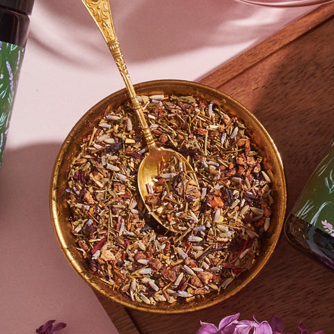 A brass bowl filled with a colorful mixture of dried herbs and spices, reminiscent of loose leaf tea, is accompanied by an ornate brass spoon. The bowl is set on a wooden surface with a bottle of Magic Hour's Blueberry Lavender Mint: Cosmic Garden Iced Tea featuring floral designs partially visible to the left.