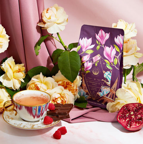 An elegant setup featuring a cup of tea on a saucer, a few pieces of chocolate, raspberries, half of a pomegranate, and a purple bag of **Breakfast in Paris-Oolong Black Tea Scented with Fruits & Flowers of Secret Parisian Gardens** adorned with floral designs. The scene evokes the charm of mystic Paris, decorated with yellow and pink flowers and luxurious fabric in the background.