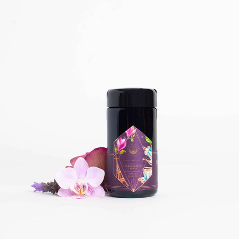 A dark-colored cylindrical container of "Breakfast in Paris-Oolong Black Tea Scented with Fruits & Flowers of Secret Parisian Gardens" sits against a white background. The container features a colorful geometric label with botanical designs reminiscent of Parisian gardens. Beside it is an arrangement of flowers, including a pink orchid and a red rose, adding a touch of elegance.