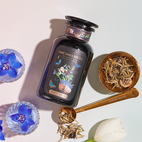 A glass jar labeled "Magic Hour Butterfly Blue Coconut Cream Iced White Tea" is surrounded by two glass cups with blue flowers, a gold spoon with loose leaf tea, and a white tulip. The golden spoon is engraved with "Magic Hour," evoking the perfect moment for summer hydration. The scene is lit with soft, natural light.