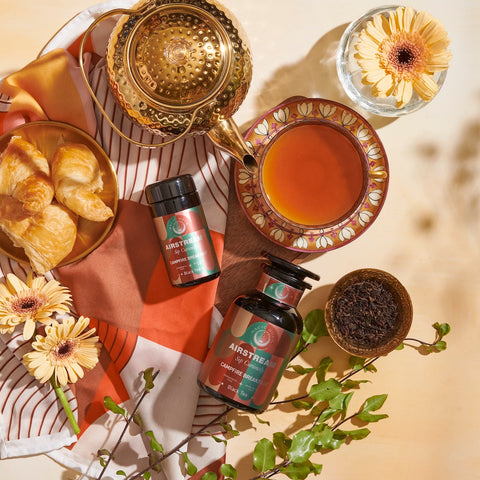 A warm scene featuring Campfire Breakfast Loose Leaf Tea from Magic Hour with teacups, a teapot, flowers, and pastries on a striped cloth. A small bowl of loose leaf tea and a vibrant leafy branch add a touch of nature to the setup. The aroma of Campfire Breakfast Loose Leaf Tea fills the air, making the atmosphere cozy and inviting.