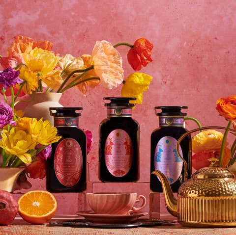 Three ornate bottles with colorful labels are placed on a shelf amidst vibrant flowers. In the foreground, a teapot and teacup set ready for Year of the Rabbit Carrot Cake Tea sits alongside a sliced orange. The pink textured wall in the background gives the scene a warm and inviting feel, perfect for a caffeine-free moment.