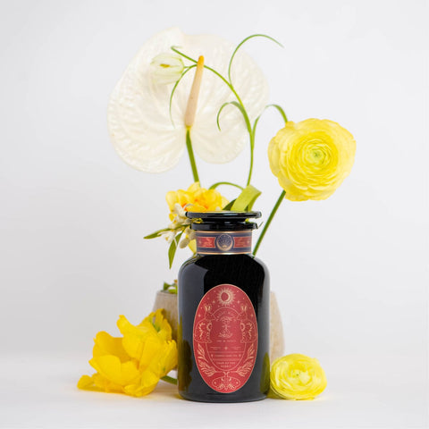 A decorative jar with a red label sits in front of a floral arrangement featuring white anthurium and yellow ranunculus flowers. The dark, ornately designed jar, labeled "Year of the Rabbit Carrot Cake Tea," holds an herbal tea. The flowers add a bright and cheerful element to the minimalist white background.