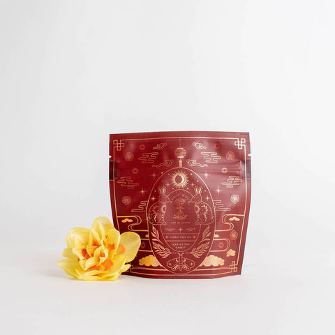 The Year of the Rabbit Carrot Cake Tea comes in a red-brown resealable bag adorned with decorative gold designs, including two rabbits and celestial motifs representing the Chinese Zodiac sign. The bag stands upright, and to its left is a yellow flower with delicate petals, both set against a plain white background.