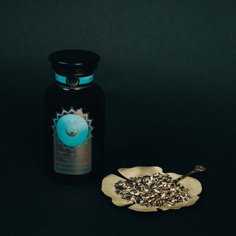 A dark glass jar with a blue and brown label labeled "Quintessence™ Tea for Opening & Healing the Throat Chakra" by Magic Hour sits next to a flower-shaped dish filled with a dried herbal mixture of organic loose leaf tea. A small spoon rests on the dish, all set against a dark background.