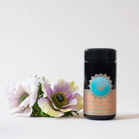 A black cylindrical container of Quintessence™ Tea for Opening & Healing the Throat Chakra with a pink and blue label sits next to a small bouquet of pale pink and purple flowers. The label shows the brand name "Magic Hour" and other text, with a turquoise emblem at the top. The background is plain white.