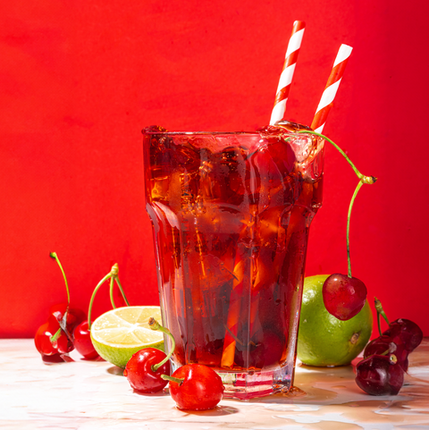 A glass of iced Magic Hour Cherry Cola with two striped straws, garnished with fresh cherries, a lime half, and a whole lime. The drink is placed against a vibrant red background, with some cherries and lime arranged around the glass.