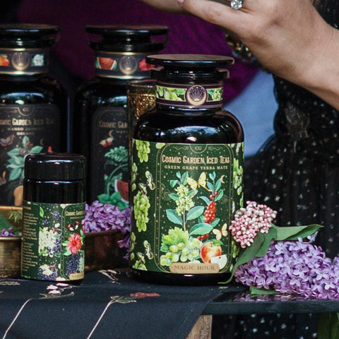 Close-up of various Magic Hour tea products adorned with vibrant floral and fruit illustrations. The focal point is a large jar labeled "Green Grape Yerba Mate: Cosmic Garden Iced Tea." Nearby are fresh lilac blooms, a delicately manicured hand, and an elegant tin of organic loose leaf tea.