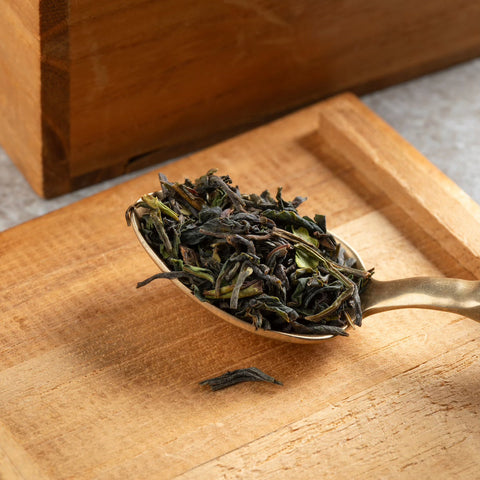 A close-up shot of a gold-colored spoon filled with April 2024 Harvest - Grand Cru First Flush Organic "Spring Wonder" Samabeong Estate Darjeeling STGFOP1 from Magic Hour. The spoon rests on a wooden surface with a slightly textured, light brown finish. In the background, part of a wooden box is visible. The organic tea leaves are dark green and twisted.