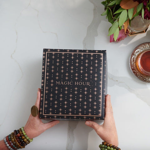 A pair of hands, adorned with beaded bracelets, holds a black box with gold star patterns and the words "Branded Gift Box" on it. The hands rest on a marble surface beside a cup of Magic Hour Tea on a decorative saucer and a small green plant with red leaves.