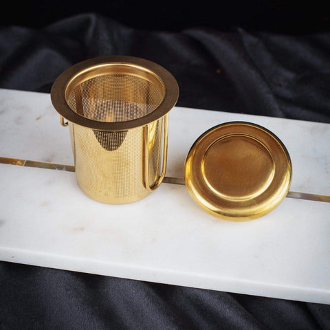 A gold-colored Midas Touch: Golden-Hued Tea Strainer by PMB Mesh Screen Company with fine mesh and a matching lid is displayed on a white marble surface with a dark fabric backdrop. Perfect for steeping your favorite loose leaf tea, the strainer stands upright, with the lid placed beside it.