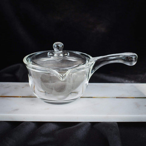 A small transparent glass teapot, the Tea-in-Hand: The Perfect Steep Side-pour Ceremonial Teapot by Espresso Parts, with a lid and a metal strainer inside, sits on a rectangular white marble slab. The background is dark and fabric-like, emphasizing the teapot's clear material and simple design, perfect for steeping your favorite loose leaf tea.