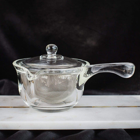 A small clear glass teapot with a lid and spout, featuring a metal mesh infuser inside, is placed on a white surface against a dark background. Perfect for brewing Magic Hour Tea, the Tea-in-Hand: The Perfect Steep Side-pour Ceremonial Teapot by Espresso Parts has a handle on one side and appears elegant and minimalist.