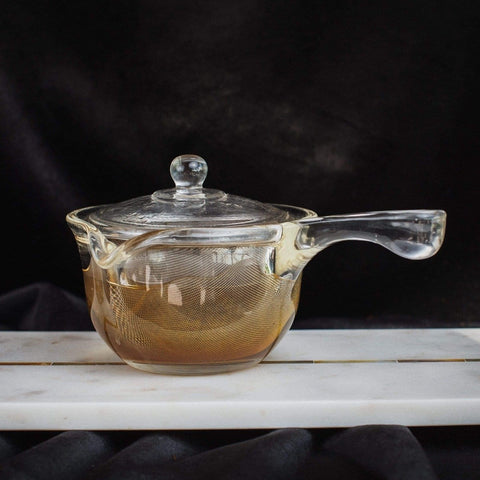 A transparent glass teapot with a long handle and a round lid is placed on a white rectangular marble base against a dark background. The Espresso Parts Tea-in-Hand: The Perfect Steep Side-pour Ceremonial Teapot, containing Organic Tea, reveals a light brown liquid with a mesh strainer visible inside.