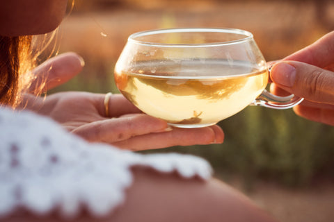 A close-up of a person holding a small, clear glass cup filled with light-colored Ti Quan Yin Oolong: Tea of Patient Compassion by Magic Hour. Another person's hand is gently holding the cup's handle. The person in the foreground is wearing a white, lace outfit, and the background is blurred with warm, natural colors.