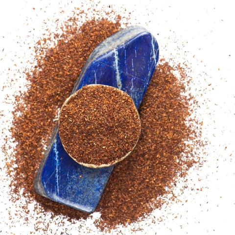 A circular pile of finely ground Rise & Shine Herbal Tea leaves from Club Magic Hour is placed on a blue stone. The blue stone serves as a base, with the tea leaves evenly spread over it and scattered around, creating an earthy and textured visual against a white background.