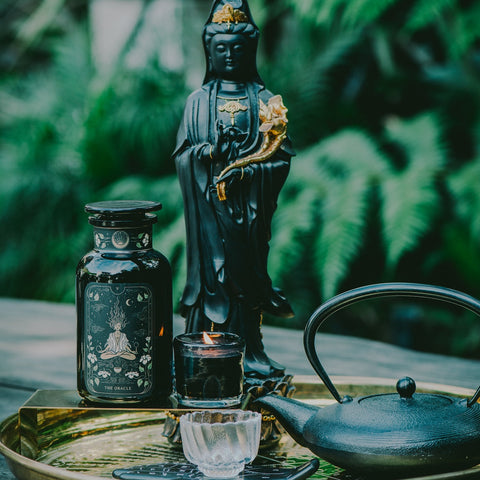 A tranquil arrangement showcases a black statue holding a scroll, accompanied by a black jar adorned with intricate designs, and a lit black candle placed in front of the statue. Next to them sits The Oracle: Lemon Lavender Tea for Supporting the Gut-Brain Axis Case Packs from Magic Hour, steeped in a cast iron teapot and poured into a small glass cup on a golden tray. The serene setting is complemented by greenery in the background, evoking an outdoor oasis.