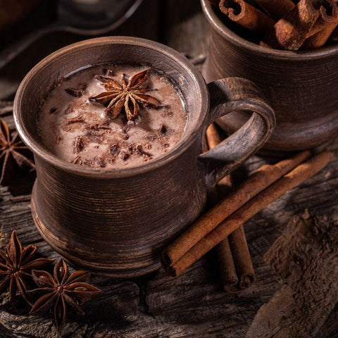 A rustic wooden mug filled with Queen of the South: Delicious Cocoa Detox Tea, garnished with star anise and chocolate shavings. Next to the mug are cinnamon sticks and another mug filled with whole cinnamon sticks, set on an aged wooden surface, creating a cozy ambiance worthy of a Magic Hour experience.