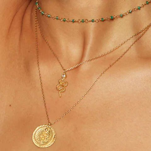 A close-up of a person wearing three layered necklaces: the top choker adorned with small green beads, the middle 18kt gold plate necklace showcasing the Enduring Individuality White Topaz Snake Necklace with a serpent symbol and white topaz detail, and the longest necklace displaying a round gold medallion with an intricate design.