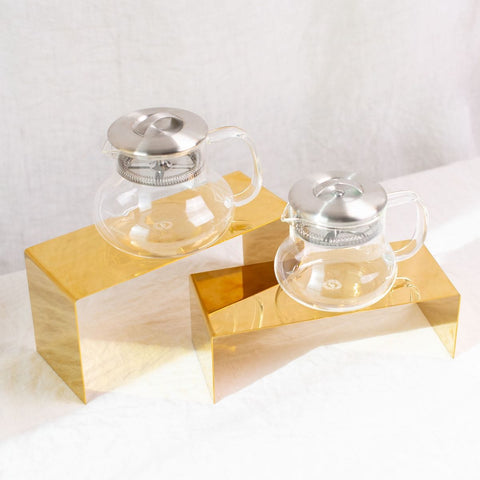 Two Essential Perfection Teapots, each featuring a built-in strainer and stainless steel lid, are displayed on gold-colored rectangular stands. Crafted from Borosilicate glass, they sit against a soft white fabric backdrop. Their simple, modern design exudes a minimalist and elegant aesthetic ideal for brewing loose leaf tea.