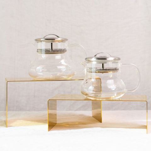 Two Essential Perfection Teapots with Built-in Strainers and metal lids are displayed on two yellow rectangular stands of different heights. These teapots, featuring built-in strainers, have a simple, modern design with round bodies, curved handles, and small spouts—perfect for enjoying loose leaf tea against the plain white fabric background.