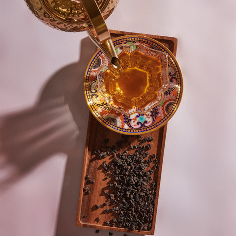 A gold teapot is pouring Magic Hour Gamma-Aminobutyric Acid (GABA) Oolong Tea into an intricately patterned, wide-mouthed glass cup placed on a decorative saucer. The cup and saucer sit atop a wooden tray scattered with loose tea leaves. This serene scene, set against a bright, clean background, offers calm nourishment for the soul.