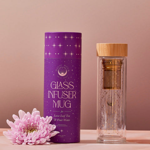 An Alibaba Glass Infuser Mug with a bamboo lid stands next to its purple cylindrical packaging labeled "Glass Infuser Mug for Loose Leaf Tea & Fruit Water." The detachable infuser and double-walled borosilicate glass ensure durability. A pale pink chrysanthemum flower is placed at the base of the packaging, all set against a neutral background.