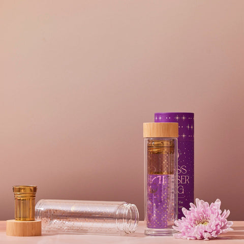 A clear glass infuser mug complete with a wooden lid and gold star accents, sits beside its star-adorned cylindrical purple packaging. The Glass Infuser Mug also boasts a detachable infuser inside. To the right of the mug, a pink chrysanthemum flower rests on the surface.