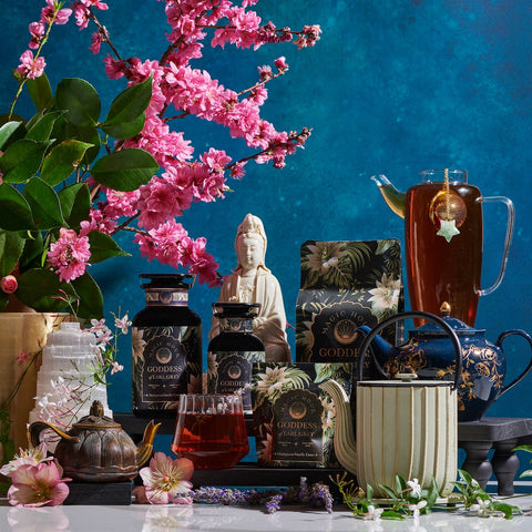 A colorful arrangement featuring a variety of organic tea products displayed on a table. The scene includes teapots, tea canisters, packets of Magic Hour's Goddess of Earl: Madagascar Vanilla Creme Tea for Soothing Delight & Delicious Decadence, a glass of tea, and a glass pitcher of iced tea with a star-shaped tea infuser. Pink cherry blossoms and a Buddha statue adorn the background.