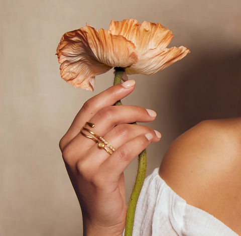 A person with manicured nails holds a delicate, light-orange flower by its stem. The person is wearing a white, off-shoulder top, and two gold rings adorned with small pearls are visible on their fingers against a soft, neutral background. One of the rings is the Gold Coils Black Snake Adjustable Ring, featuring an elegantly coiled serpent wrapped around the finger.