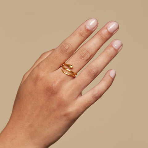 The Gold Coils Black Snake Adjustable Ring, featuring a sleek and coiled design, is elegantly worn on the middle finger against a neutral beige backdrop. The fingernails are perfectly manicured with a light pink polish.