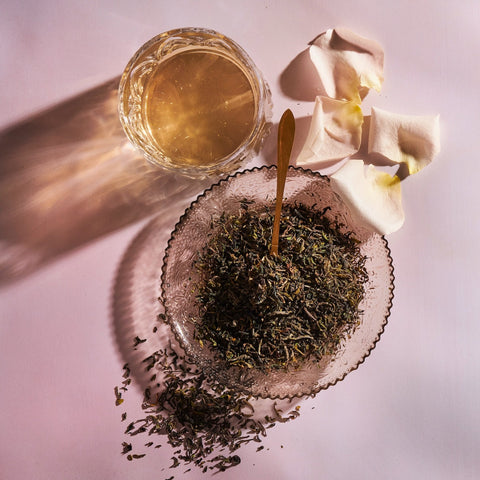 A glass of light-colored tea sits on a table near a glass bowl filled with loose Magic Hour Goomtee Estate Darjeeling tea leaves and a gold teaspoon. Three white flower petals with yellow centers are arranged around the bowl. The setup casts soft shadows on the pinkish surface, evoking the charm of Muscatel Valley hills.