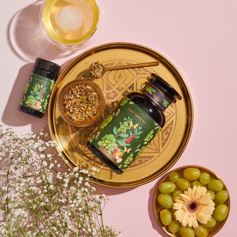 A golden tray holds a green bottle and a small jar with botanical labels, a bowl of loose leaf tea, and a golden spoon. Next to the tray are a bunch of grapes, a gerbera daisy, white flowers, and a glass with a round ice cube on a pink background. Perfect for your Magic Hour Green Grape Yerba Mate: Cosmic Garden Iced Tea experience.