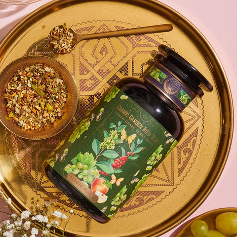 A decorative gold tray holds a dark green bottle of organic tea labeled "Green Grape Yerba Mate: Cosmic Garden Iced Tea" by Magic Hour, with images of flowers, herbs, and fruits. The tray also has a small wooden bowl filled with loose tea leaves and a gold spoon resting beside it.