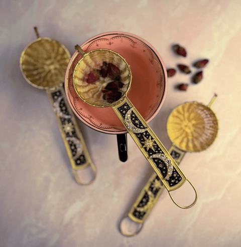 A 22-Carat Gold Plated Radiant Moon Gemstone Tea Strainer from Jaipur, India, filled with rose petals, rests on the rim of a pink teacup. The gold-plated strainer features intricate black and gold designs. Two additional brass tea baskets lie flat on the marble surface beside scattered dried rose petals.
