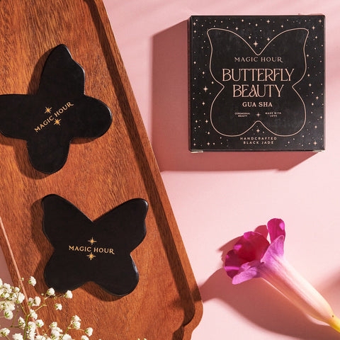 A flatlay showcasing butterfly-shaped black jade Butterfly Beauty Gua Sha tools by Magic Hour, one on a wooden tray and one beside a boxed set labeled "Magic Hour Butterfly Beauty." A single pink calla lily and white flowers accent the pink background, evoking the serene elegance of Magic Hour Tea.