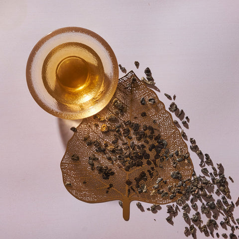 A glass cup filled with a light amber liquid sits on a light pink surface next to a golden leaf-shaped tray. Loose Magic Hour Gunpowder Green Tea leaves are scattered from the tray onto the surface, adding an earthy flavor to the scene. The arrangement creates an aesthetically pleasing and artistic presentation.