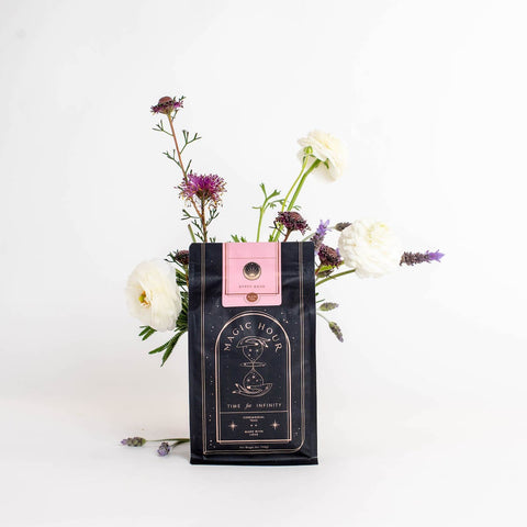 A black and pink package of "Gypsy Rose Black Tea" by Magic Hour stands on a white surface, adorned with the label "Time for Infinity". A few white and purple flowers, including lavender, are creatively arranged around and behind the package, giving it a fresh, elegant look.