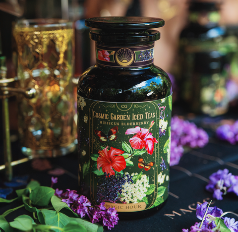 A decorative bottle of Magic Hour Hibiscus Elderberry: Cosmic Garden Iced Tea sits on a table surrounded by purple flowers. The label, showcasing vibrant illustrations of hibiscus flowers, elderberries, butterflies, and foliage, gives it a magical and whimsical appearance reminiscent of Organic Tea at Magic Hour.