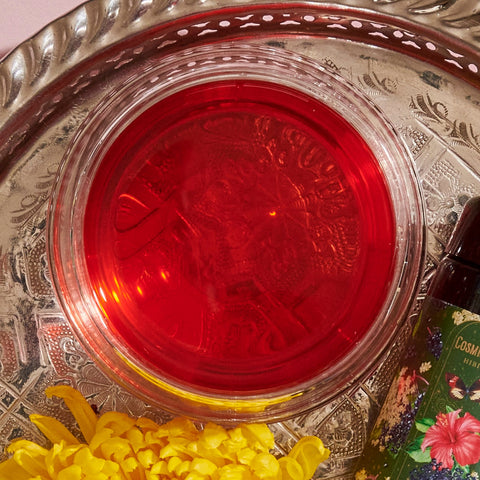 A round glass bowl filled with a bright red liquid is placed on an ornate silver tray. Beside the bowl, a yellow marigold flower and a colorful bottle with a floral design are visible, hinting at Hibiscus Elderberry: Cosmic Garden Iced Tea by Magic Hour. The setup appears decorative and festive.