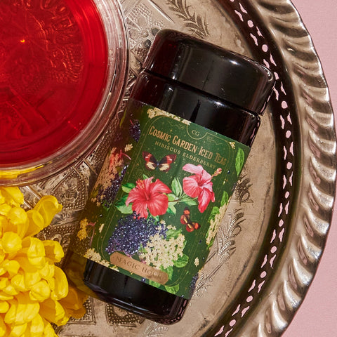 A tin labeled "Hibiscus Elderberry: Cosmic Garden Iced Tea" with floral artwork sits on an ornate silver tray. To its left is a glass of red liquid, and to its right are yellow flowers. The background features a soft pink surface, perfectly showcasing the allure of Magic Hour.