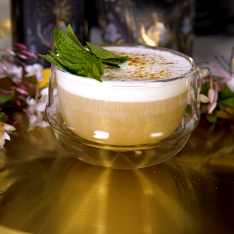 A glass cup filled with The High Priestess: Wisdom Tea for Powerful Dreams & Illuminated Insights by Magic Hour, garnished with fresh mint leaves on top, sits on a golden surface. In the background, there are floral elements and ornate black and gold decorative items.