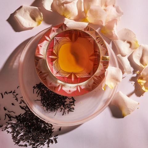 A cup of Idulgashinna Black Tea in an ornate pink and gold teacup and saucer sits on a light surface. Tea leaves from the Idulgashinna Tea Estate are scattered next to the cup along with white rose petals. Sunlight creates soft shadows, adding a warm ambiance to the scene.