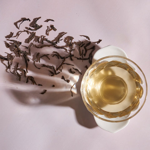 A clear glass cup filled with light-colored Ceylon Biodynamic Green Tea sits on a white saucer. To the left of the cup, scattered dried tea leaves cast shadows on a light pink surface. The scene is lit from the top left, creating a tranquil and minimalistic composition.