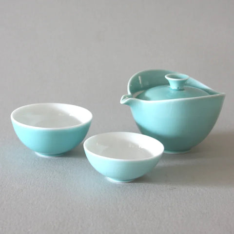 The Ingot Blue Travel Tea Set from Eastern Elm includes a teapot with a lid and two small teacups set against a light gray background. The cups and teapot feature a harmonious color palette, combining soft teal hues with white interiors. The minimalist and elegant design makes it ideal for portable enjoyment.